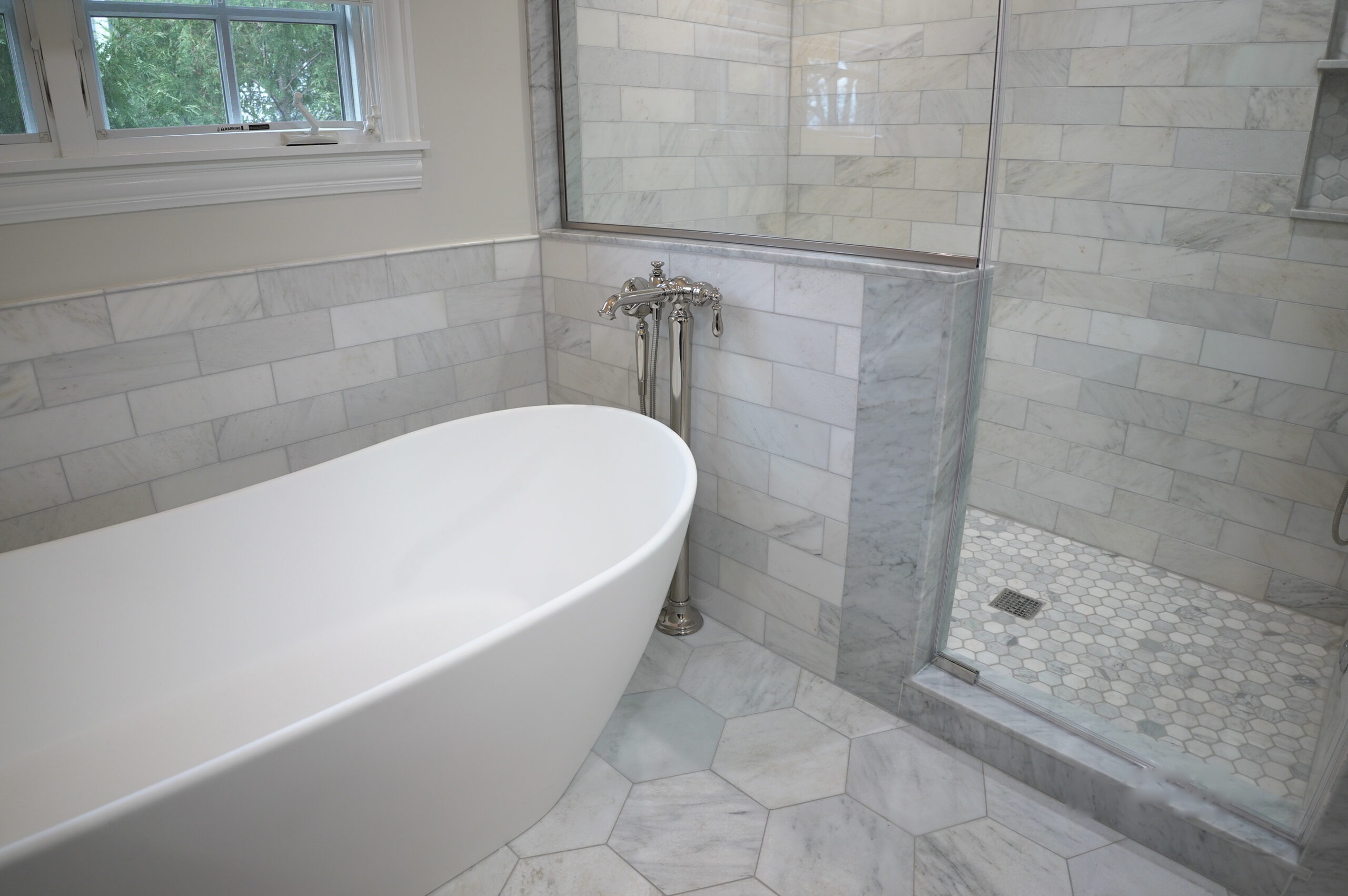 Freestanding soaking tub with a floor-mounted nickel filler beside a glass-enclosed shower, marble subway tile walls and a hexagon marble floor